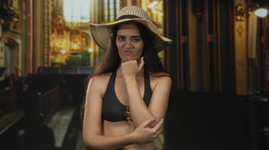 Kilise binasında siyah bikinili genç bir kadın ve eli çenesinde hasır şapka giyiyor..