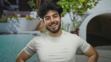 Sakallı genç bir adam otel ortamında havuz kenarında gülümsüyor, yemyeşil ve modern mimarinin ortasında rahat bir tatil havası taşıyor..