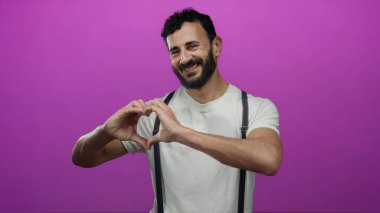 Sakallı, kalp şeklinde, elleri pembe arka planda olan İspanyol bir adam. Askılı, sıradan beyaz bir gömlek içinde mutluluk ve aşk yayıyor..