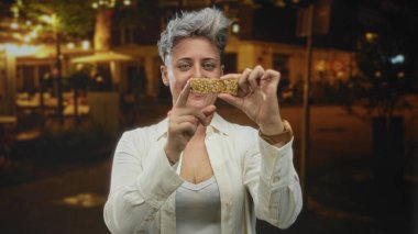 Gece, elinde granola çubuğuyla gülümseyen ve kentin caddesinde atıştırmak için parmağını gösteren bir kadın. Mutluluk..