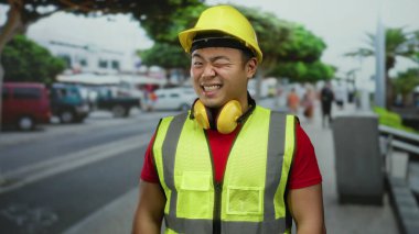 Güvenlik teçhizatı ve sert şapkalı güler yüzlü adam şehir yaşamının ortasında inşaat sektöründe profesyonelliğini sergileyen kalabalık bir caddede kendinden emin duruyor..