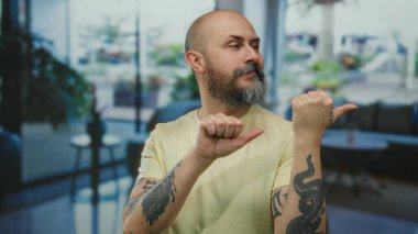 Sakallı ve dövmeli kel bir adam otel ortamında içeriyi işaret ediyor, rahat ve davetkar bir atmosferi taşıyor..