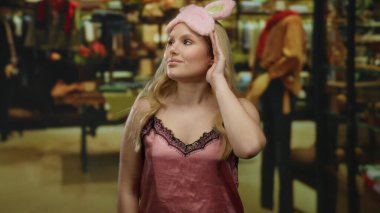 Pembe uyku maskeli sarışın kadın bir giyim mağazasında duruyor, şık gösteriler ve renkli giysiler arasında merak uyandırıyor..
