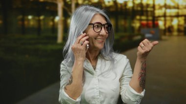 Gri saçlı ve gözlüklü bir kadın telefon pazarının dışında konuşurken başparmağıyla kenarı işaret ediyor, sıradan beyaz bir gömlek giyiyor ve kollarında dövmeler gösteriyor..
