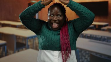 Sınıfta neşeli hareketler yapan bir kadın, elleri başının üstünde, pembe örgüleri yeşil süveterin altında, heyecanını ya da eğlencesini ifade ediyor..