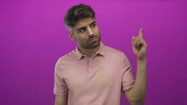 Sıradan pembe tişörtlü genç İspanyol adam mor stüdyo ortamındaki tavanı işaret ediyor..