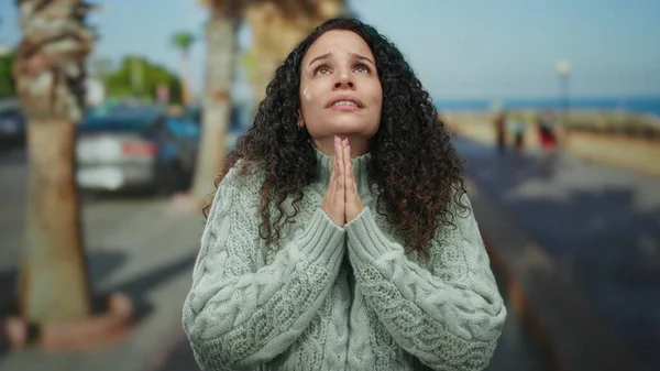 Arabaları ve palmiye ağaçları olan bir sokakta, güneşli bir günde, mavi gökyüzünün altında umut dolu ve huzurlu görünen, rahat bir kazakla dua eden bir kadın..