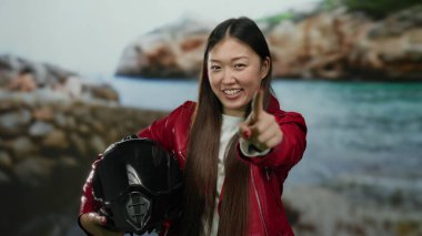 Sahilde kayalık arka planda kaskını tutan kırmızı ceketli bir kadın gülümsüyor..