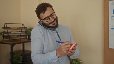 İspanyol bir adam, modern ofis ortamında birden fazla iş yaparken notlar yazıyor. Kulağının ve omzunun arasında bir telefon tutuyor. Yoğun ama odaklanmış bir çalışma ortamı yaratıyor..