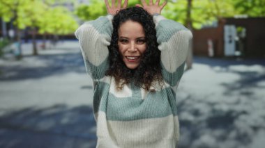 Genç bir kadın, güneşli bir günde arka planda ağaçlarla dışarıda dikilirken elleri kafasında tavşan kulağı hareketi yapıyor..