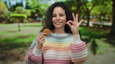 Parkta elinde kurabiyeyle gezen, renkli kazaklı, kıvırcık saçlı ve güneşli bir günde gülümseyen bir kadın..