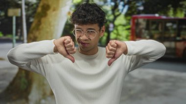 Gözlüklü ve beyaz kazaklı bir adam iki eli sokakta baş parmak hareketi gösteriyor. Onaylanmamış tepki.