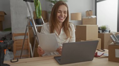 Taşınan kutularla dolu binada elinde dizüstü bilgisayarla belgeyi tutan kadın yumruğunu sıktı; başarı için iyimserlik, dayanıklılık ve neşe.