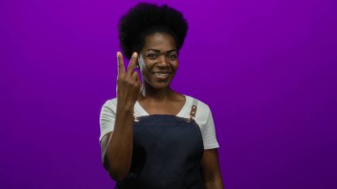 Önlükle gülümseyen bir kadın canlı mor arka planda neşeli bir ifadeyle pozitif bir hareket sergiliyor..