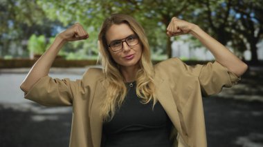 Güneşli bir parkta güvenle duran, kollarını esneten, gözlüklü ve parlak yeşil ağaçlarla çevrili bir spor ceket giyen bir kadın..