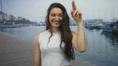 Uzun saçlı genç İspanyol kadın güneşli bir günde deniz kenarındaki bir rıhtıma selam veriyor ve gülümsüyor..