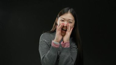 Siyah arka planda sıcak süveter giyen, uzun saçlı, neşeli yüzlü, neşeli bir kadın tezahürat ve coşkuyla bağırıyor..