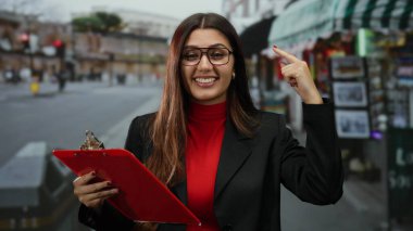Gözlüklü genç bir kadın işlek caddede kırmızı pano tutarken gülümsüyor ve bulutlu gökyüzünün altında kafasını işaret ederek canlı şehir enerjisi ve özgüvenini yakalıyor.