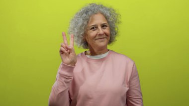 Gri saçlı, gülümseyen ve hayat dolu sarı bir arka plana barış işareti koyan pembe kazaklı, mutluluk ve pozitiflik taşıyan bir kadın..