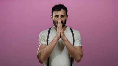Meditasyonda sakallı İspanyol bir adam pembe arka planda poz veriyor, sakin ve içe dönük bir şekilde dua ediyor, beyaz gömlek ve pantolon askısı giyiyor..
