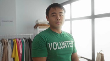 Bir hayır kurumunda gülümseyen genç bir adam yeşil gönüllü tişörtü giyip arka planda giyinmiş, toplum ruhunu ve hizmeti somutlaştırıyor..