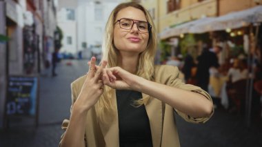 Şehir caddesindeki terasta gözlüklü, bej ceketli, gözlüklü, modern iletişim ve sosyal medya nişanını gösteren hashtag jestleri yapan bir kadın..