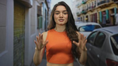 Dar şehir caddesinde kameraya doğru dik dik bakan ve avuç içi açık turuncu elbiseli kadın, sakinlik..