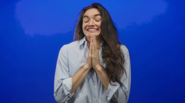 Mavi bir stüdyoda şans dileyen bir kadın gözleri kapalı gülümseyen parlak ve uzun siyah saçları omuzlarından dökülen; umut.