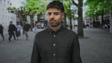 Genç Hispanik adam sıkılmış çene ve yoldan geçen yayaların ortasında çatık kaşları ile kaşlarını çatar; iğrenme.