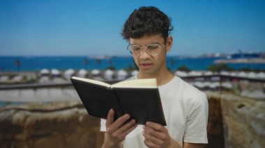 Parlak güneş ışığı altında, deniz ufkuna odaklanmış bir bakışla limanda tırabzan kullanarak ciltli kitap okuyan adam:.