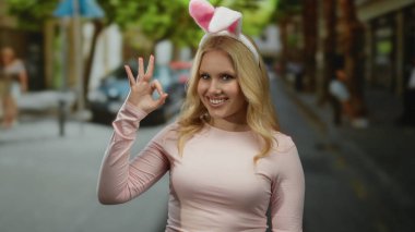 Sarı saçlı ve tavşan kulaklı gülümseyen kadın şehir sokaklarında şakacı hareketler yapıyor, açık havada neşeli şehir atmosferi sergiliyor..