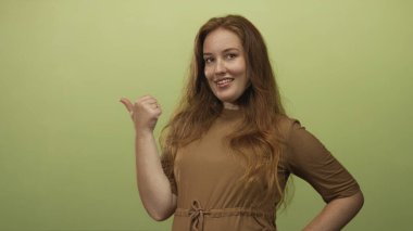 Kafkasyalı genç bir kadın gülümsüyor ve kahverengi elbise giyerken yumuşak yeşil stüdyo duvarını işaret ediyor..