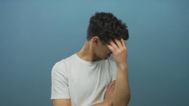 Gözlüklü genç İspanyol adam alnını tutuyor ve kollarını mavi bir stüdyo arkaplanına çarpıştırıyor; stres.