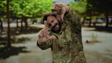Askeri üniformalı İspanyol bir adam ağaçlarla dolu bir sokakta dikilirken manzarasını elleriyle çerçeveliyor..