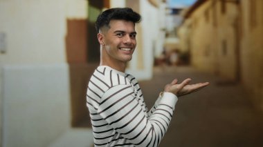 Çizgili tişörtlü genç adam elleri yana doğru uzanmış bir şekilde gülümsüyor şehir sokaklarında rahat ve mutlu bir şehir havası sergiliyor..