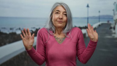 Gri saçlı kadın, deniz kenarında pembe elbiseli, bulutlu gökyüzünün altında okyanus manzaralı bir yerde rahatlama ve tefekküre dalma hareketi yapıyor..