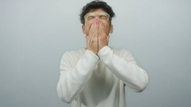 Beyaz kazaklı genç İspanyol adam, gri stüdyo ortamında sessizlik için parmaklarını dudaklarına bastırıyor..