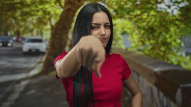 Kırmızı tişörtlü İspanyol kadın, arka planda ağaçlar ve arabalarla şehir sokaklarında öfkesini sergiliyor, güçlü duygusal ifadeyi vurguluyor..