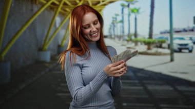 Elinde İsveç banknotları tutan bir kadın, şehir sokaklarında gülümsüyor, kızıl saçlı bir yetişkinin neşeli ifadesini sergiliyor..