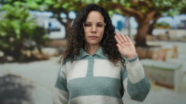 Woman waving outdoors in park wearing glasses and striped sweater surrounded by trees in a natural, relaxed setting on a sunny day portraying a casual outdoor moment.