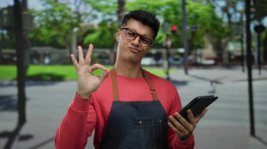 Önlüklü İspanyol bir adam şehir caddesinde tablet kullanıyor. Onay veriyor. Dışarısı canlı bir manzarayla çevrili..