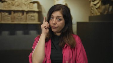 Pembe ceketli kadın, müze galerisinde parmağını parmağıyla şakağına kaldırıyor ve ciddi bir ifadeyle yansıtıyor..