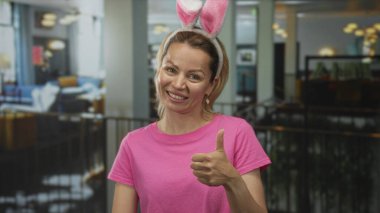 Pembe tişörtlü bir kadın, tavşan kulaklı bir kadın ve otel lobisinde sıcak ışık altında açık avuç içi jestleri, sıcak bir konukseverlik..