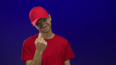 Gözlüklü, kırmızı şapkalı, gülümseyen ve mavi stüdyoyu işaret eden İspanyol genç adam..