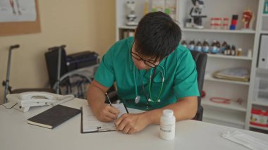 Yeşil üniformalı genç bir doktor klinikte çalışıyor, şişeyi inceliyor, not alıyor, sağlık ekipmanları ve telefon ile çevrili..