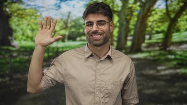 Güneşli ormanda bej gömlekli, gözlüklü, gülümseyen ve kameraya el sallayan bir adam..