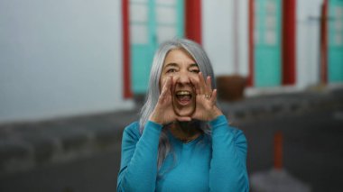 Canlı şehir ortamında renkli kapıları ve pencereleri olan canlı bir sokakta gri saçlı, mavi kazaklı yaşlı bir kadın bağırıyor..