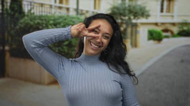 Mavi süveter giyen kadın gülümsüyor ve bina cephesinin yanında el ele barış işareti gösteriyor; gençlik neşesi.