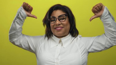 Gülümseyen, iri yarı İspanyol kadın, beyaz gömlekli, kendine güveni ve enerjisi olan, parlak sarı bir arka plana işaret ediyor..