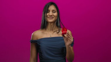 Gülümseyen kadın kırmızı yüzük kutusunu canlı pembe arka planda tutuyor. Neşe ve kutlamaları yansıtıyor. Şık, omuzsuz elbisesiyle zarafet sergiliyor..